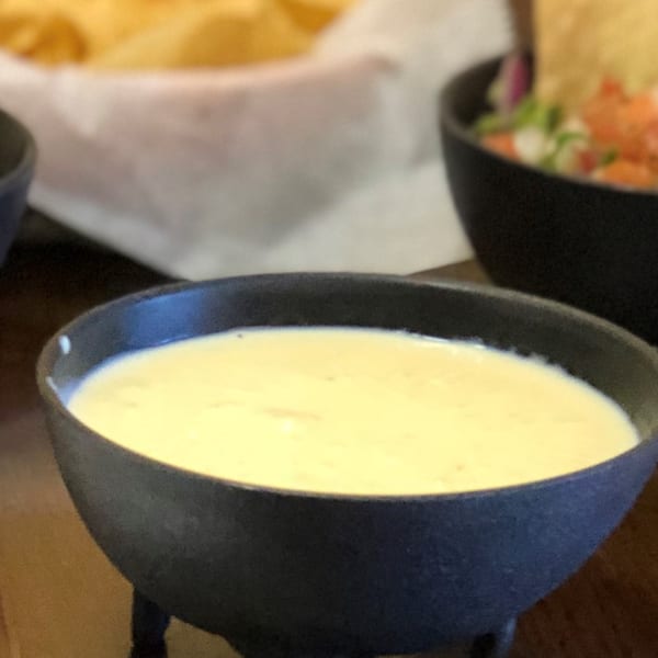 A black bowl of creamy queso dip, with blurred tortilla chips and another dish in the background.