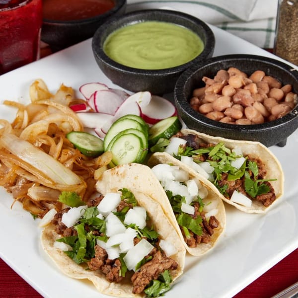 Two tacos with meat, onions, and cilantro, served with sides of beans, guacamole, and sautéed onions.
