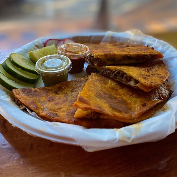 Crispy birria quesadillas with limes, cucumbers, and two sauces in a white paper-lined basket.