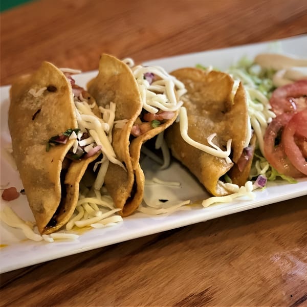 Three crispy tacos filled with meat, cheese, and vegetables, served with lettuce and sliced tomatoes.
