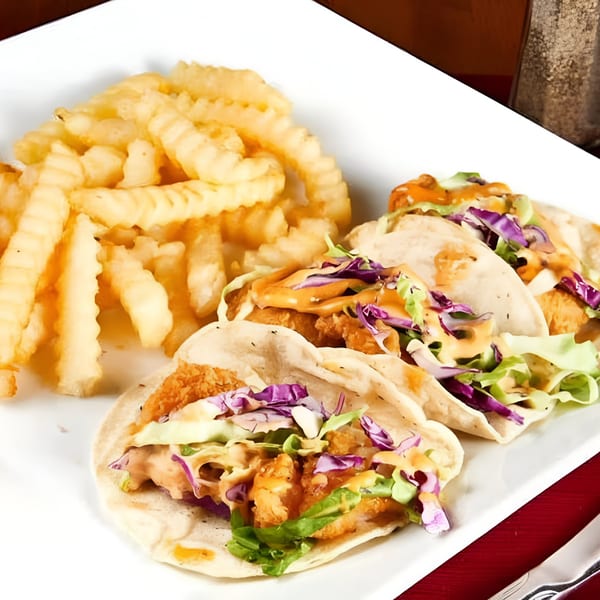 Three fish tacos with red cabbage, lettuce, and sauce, served with crinkle-cut fries on a white plate.