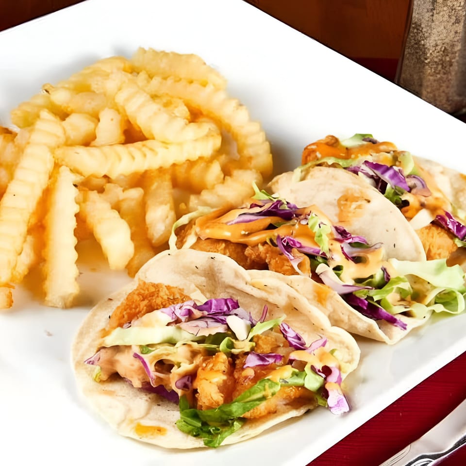 Three fish tacos with red cabbage, lettuce, and sauce, served with crinkle-cut fries on a white plate.