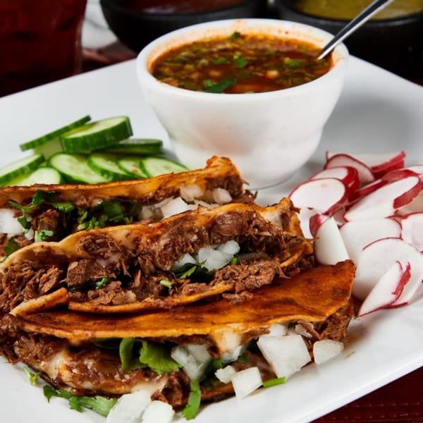 Three birria tacos with cilantro, onions, a side of consommé, sliced cucumbers, and radishes.