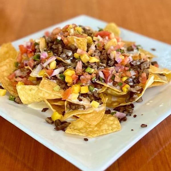 A plate of Taco-Holics Club nachos with beef, corn, tomatoes, and onions on a white plate.